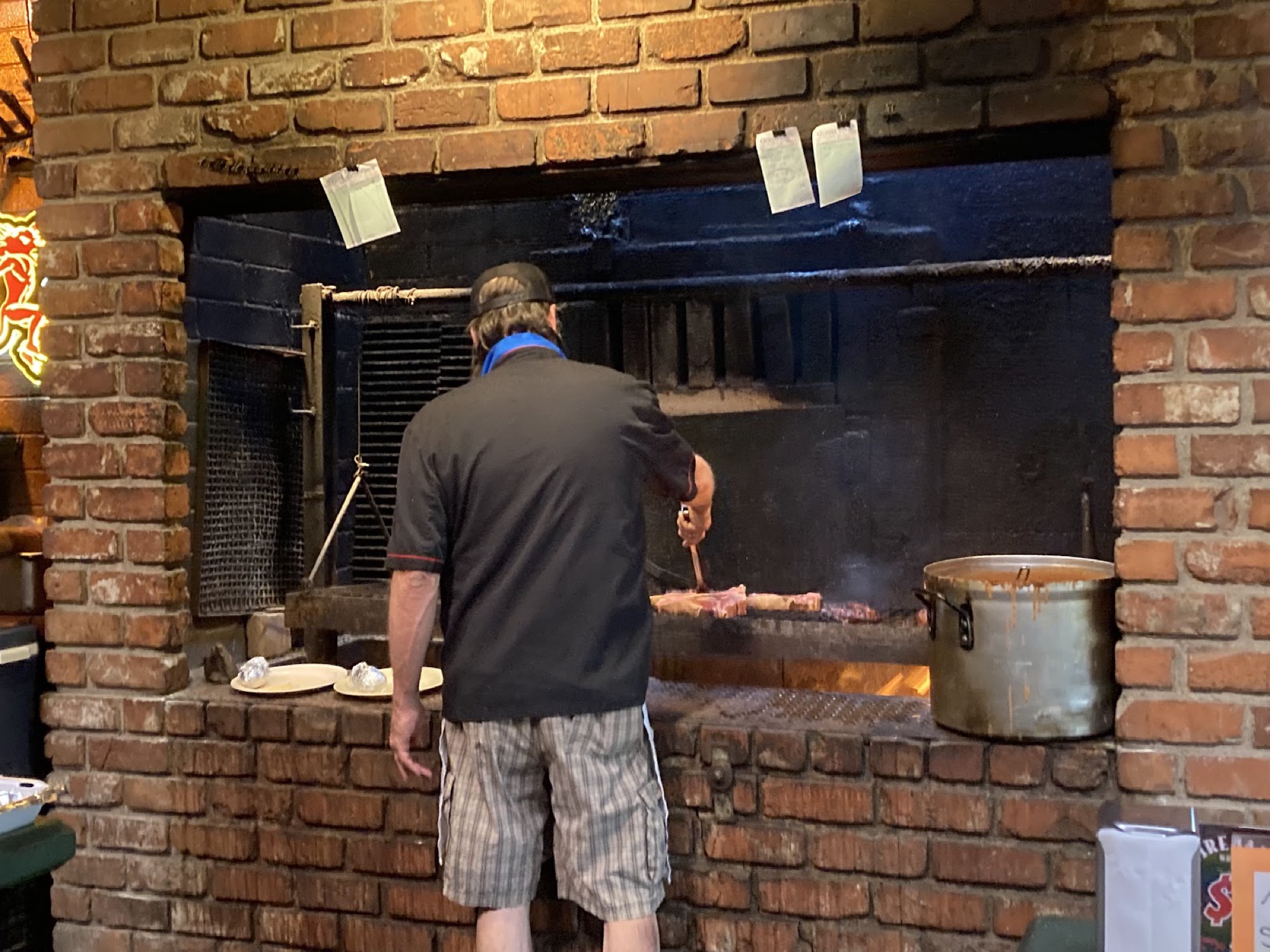 A cook tending steaks over an open brick fire pit.