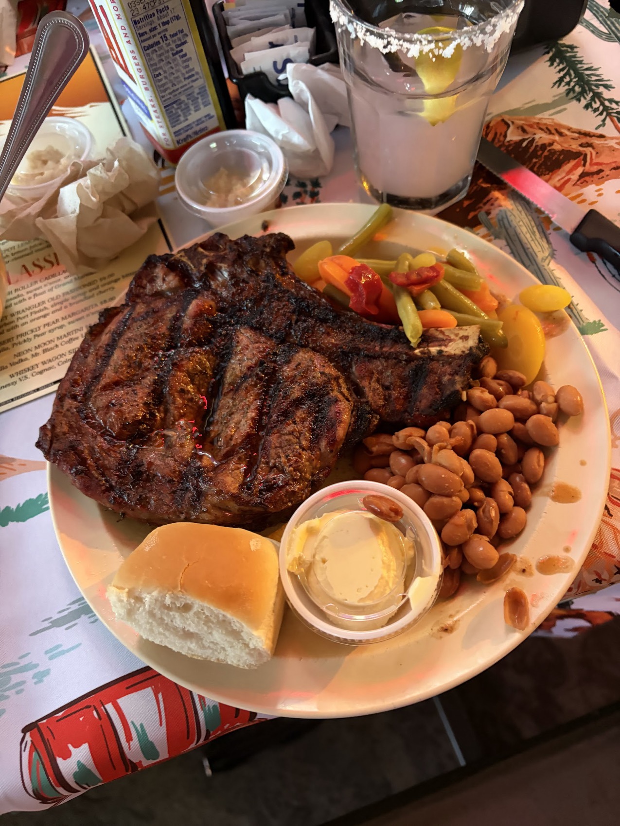 A grilled bone-in steak with beans, roasted peppers and a dinner roll.